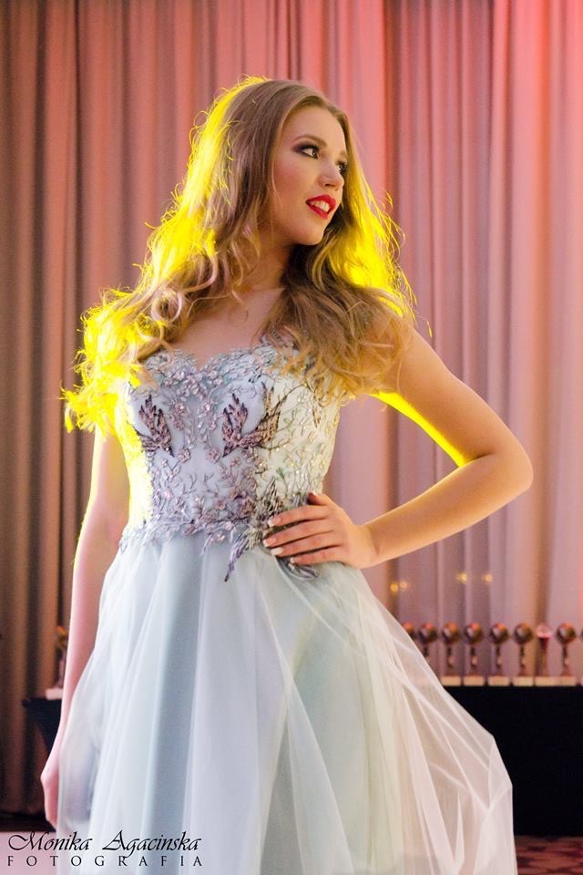 Blond modelka w błękitnej sukni balowej z haftowanymi aplikacjami na gorsecie, pozująca na tle zasłon i rzędu statuetek.