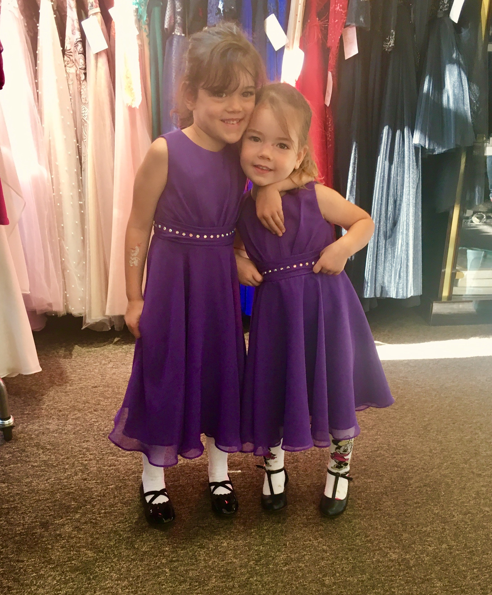 Dwie uśmiechnięte dziewczynki w fioletowych sukienkach z ozdobnym paskiem i czarnych lakierkach, jedna z tatuażem na ramieniu, pozują na tle wieszaków z sukniami.