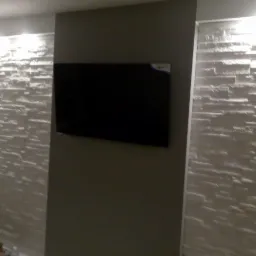 Telewizor zawieszony na szarej ścianie, otoczony dekoracyjną, białą ścianą z kamienia, oświetloną punktowo od góry.