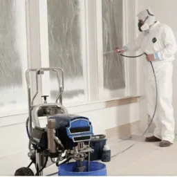 Pracownik w kombinezonie ochronnym maluje natryskowo ramy okienne na biało, używając profesjonalnego agregatu malarskiego; okna zabezpieczone folią.