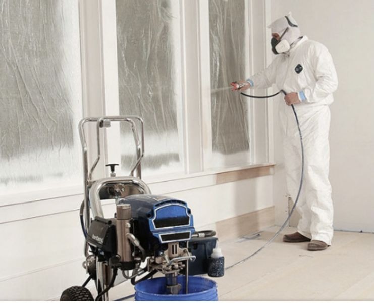 Pracownik w kombinezonie ochronnym maluje natryskowo ramy okienne na biało, używając profesjonalnego agregatu malarskiego; okna zabezpieczone folią.