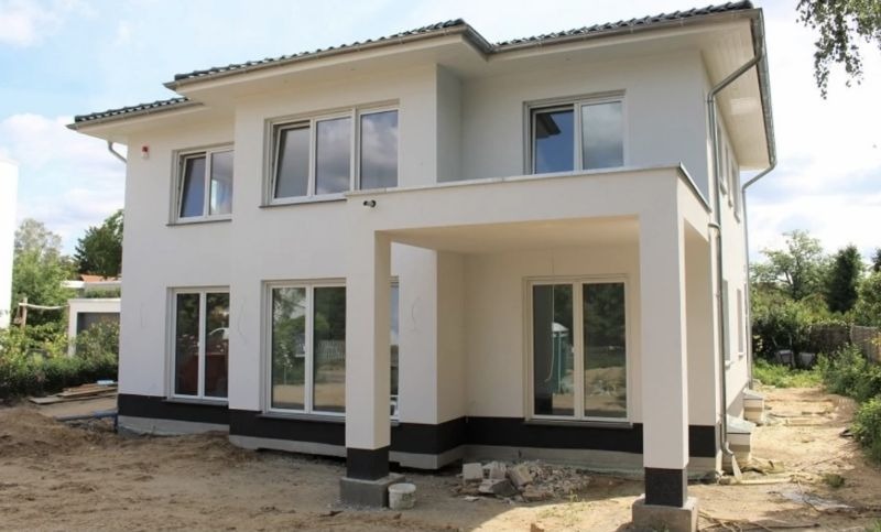 Nowoczesny, dwukondygnacyjny dom z białym tynkiem i ciemną podmurówką, widok z zewnątrz na częściowo wykończony teren.