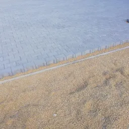 Ułożona szara kostka brukowa na podsypce z kruszywa, widoczne palety w tle i fragment budynku.