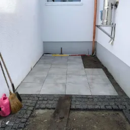 Fragment układanego tarasu: szare płyty na podłożu, obrzeże z kostki granitowej, poziomica i narzędzia przy ścianie budynku z oknem.