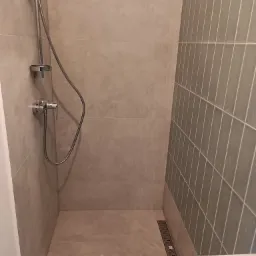 Nowoczesna kabina prysznicowa z odpływem liniowym, wykończona beżowymi płytkami wielkoformatowymi i zielonymi, wąskimi płytkami ułożonymi pionowo na jednej ze ścian.