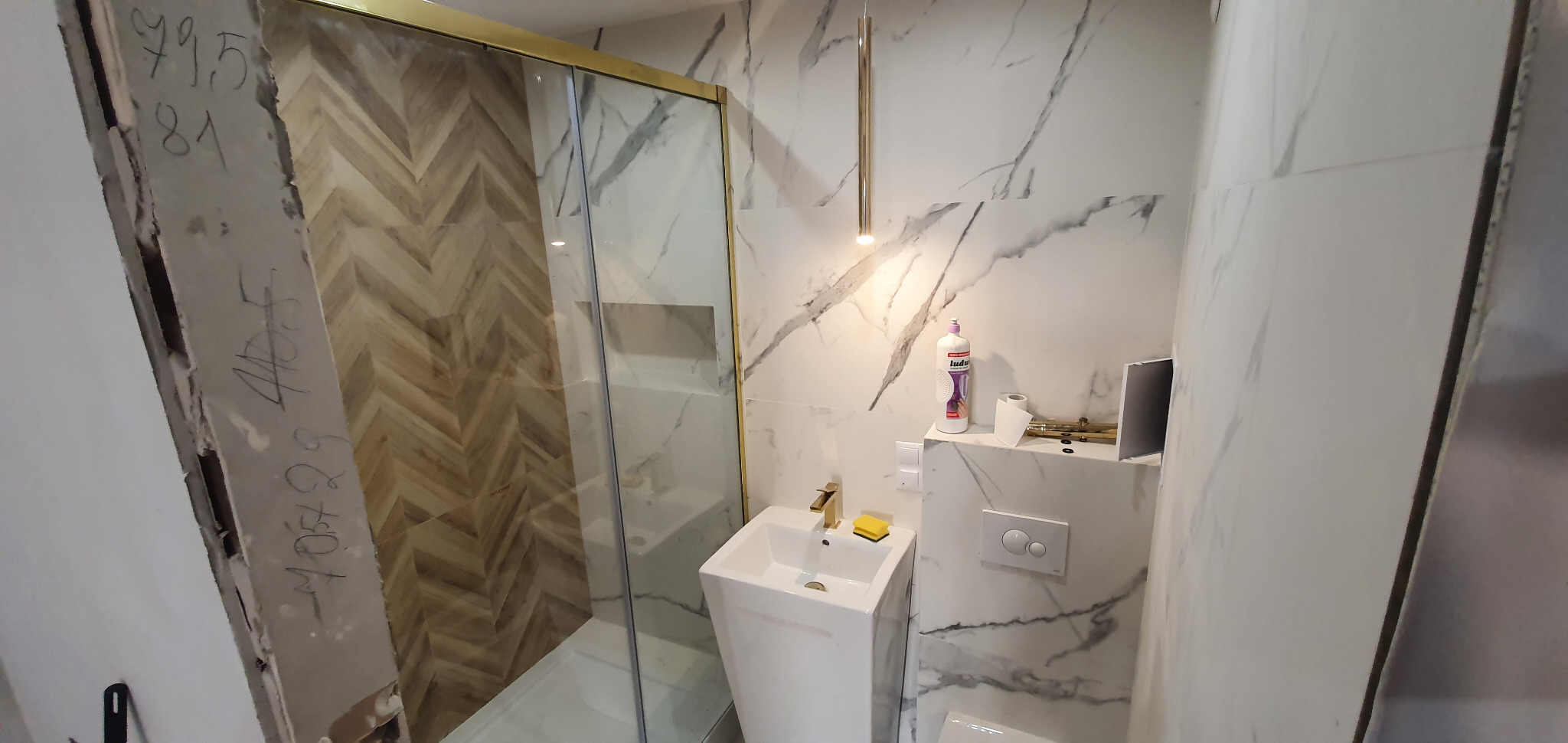 Nowoczesna łazienka z kabiną prysznicową z elementami w kolorze złotym, białą umywalką na postumencie, marmurowymi płytkami ściennymi i podłogą w jodełkę, w trakcie prac wykończeniowych, widoczne...