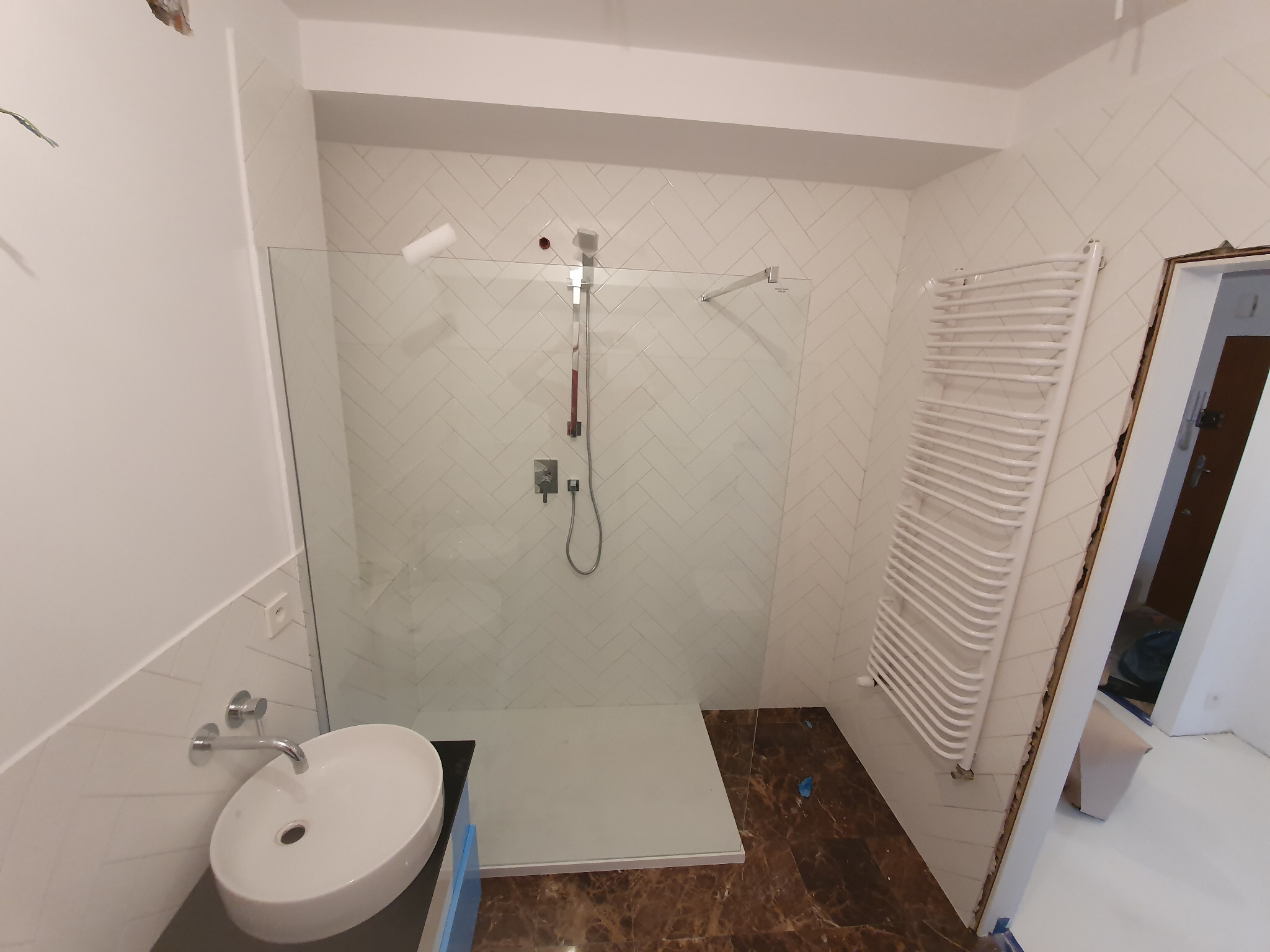 Wykończona łazienka z kabiną prysznicową typu walk-in, białymi płytkami ułożonymi w jodełkę, umywalką nablatową i grzejnikiem drabinkowym.