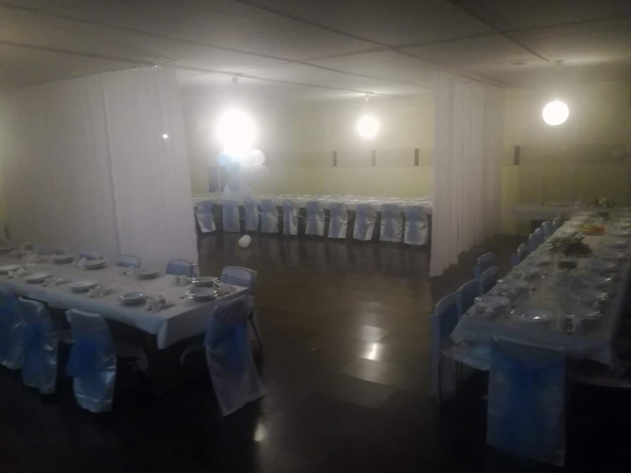 Sala bankietowa z długimi stołami nakrytymi białymi obrusami, krzesła z niebieskimi szarfami, dekoracje z białych balonów w tle, oświetlenie punktowe.