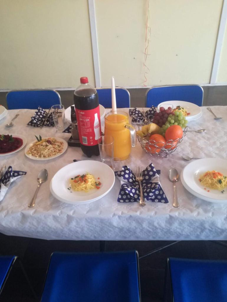 Uroczysty stół z potrawami, napojami i owocami w Dziadowej Kłodzie, nakryty białym obrusem z dekoracyjnymi serwetkami w niebieskie grochy. Widoczny dzbanek z sokiem pomarańczowym i butelka Coca-Coli.