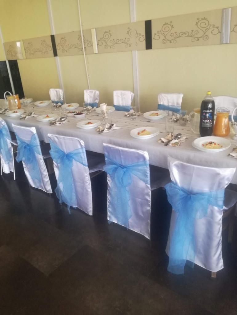 Uroczysty stół nakryty białym obrusem, białe krzesła z niebieskimi kokardami, talerze z przystawkami, napoje w dzbankach i butelkach, dekoracje na ścianie.