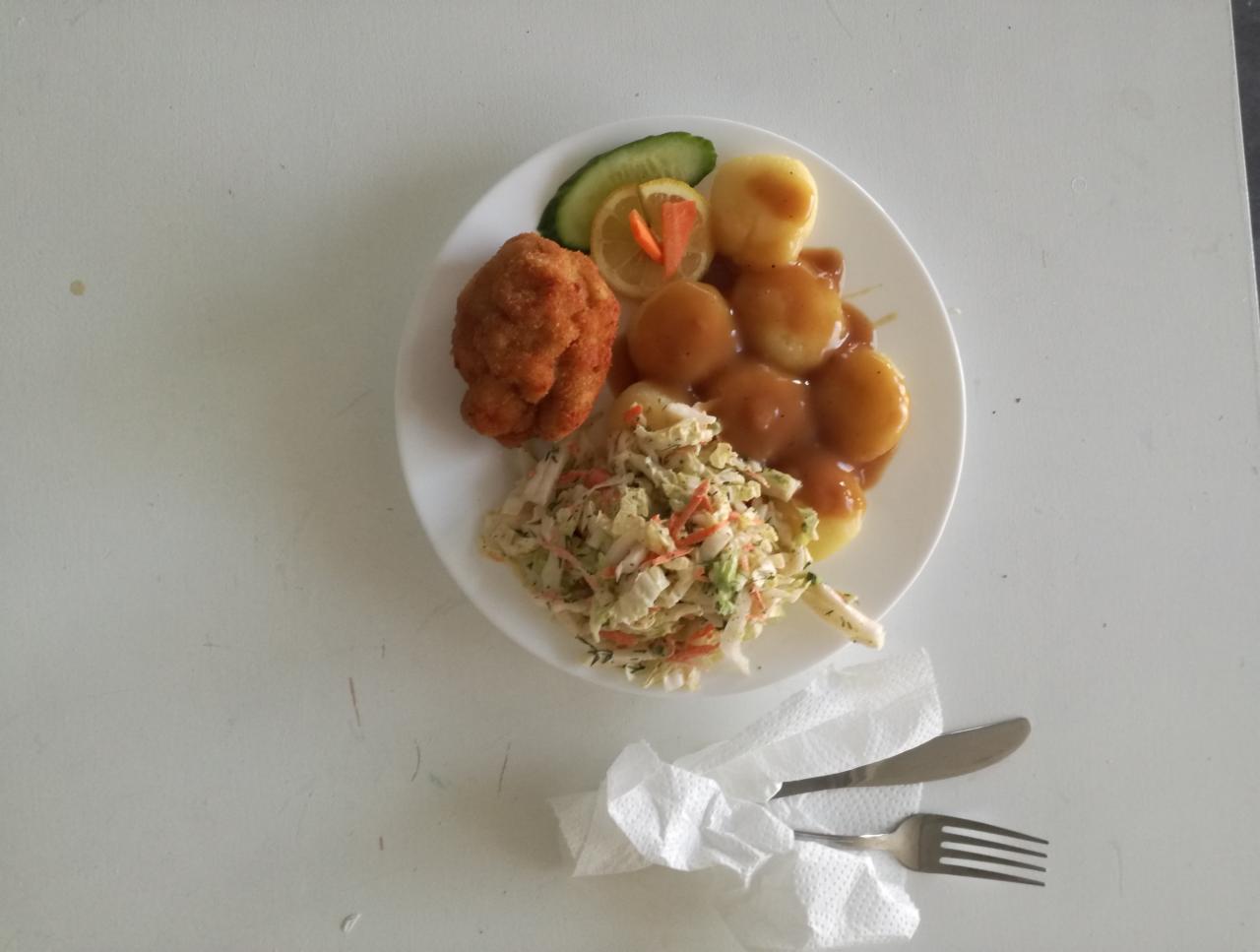Talerz z obiadem: kotlet, ziemniaki w sosie, surówka z białej kapusty, ogórek i cytryna, ułożone na białym talerzu, obok sztućce owinięte w serwetkę.