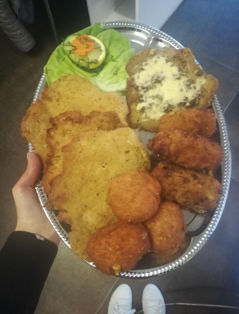 Srebrna patera z kotletami schabowymi, krokietami i kotletem z serem, udekorowana sałatą i cytryną z marchewkową dekoracją, trzymana przez osobę w czarnej bluzie i białych butach.