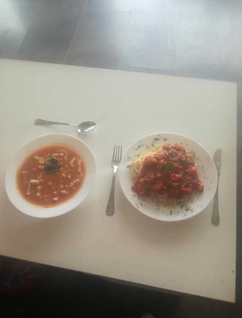 Dwa dania na białym stole: zupa pomidorowa z makaronem i spaghetti z sosem pomidorowym, sztućce obok talerzy.