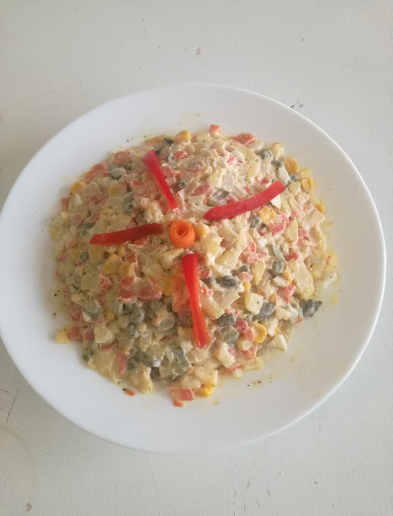 Tradycyjna sałatka jarzynowa na białym talerzu, udekorowana paskami czerwonej papryki ułożonymi w kształt promieni i rozetką z marchewki pośrodku.