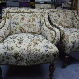 Dwa odnowione fotele w kwiatowy wzór z ciemnymi, toczonymi nogami, widoczne na tle magazynu tkanin.
