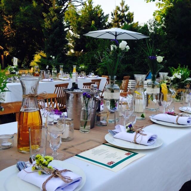 Uroczysty stół na zewnątrz z białym obrusem, srebrnym wiaderkiem na lód, szklanymi naczyniami z dekoracjami kwiatowymi i menu z zieloną obwódką.
