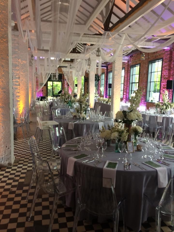 Elegancka sala bankietowa z okrągłymi stołami nakrytymi szarymi obrusami, przezroczystymi krzesłami, kwiatowymi dekoracjami i szachownicą na podłodze.