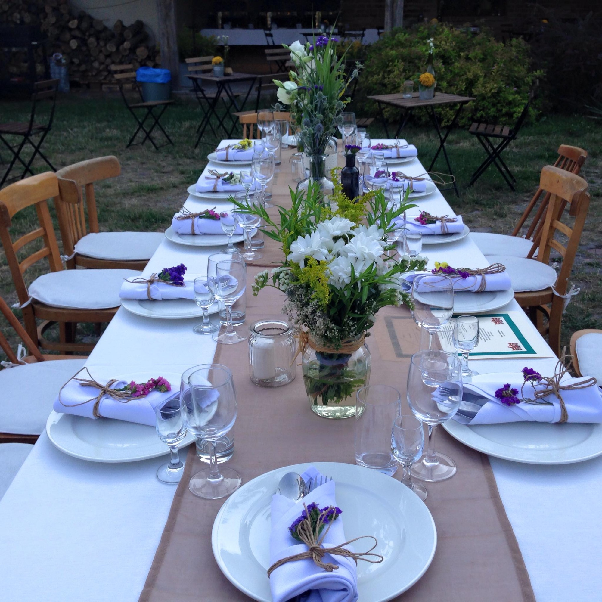 Długi stół udekorowany białym obrusem, brązowym bieżnikiem, kwiatami w wazonach, białymi talerzami z serwetkami przewiązanymi sznurkiem i fioletowymi kwiatami, szklankami oraz krzesłami w ogrodzie.