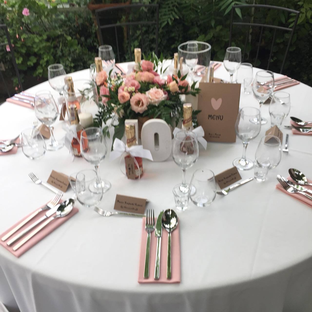 Elegancko nakryty okrągły stół z białym obrusem, różowe serwetki, srebrne sztućce, szklanki, mini butelki z alkoholem z kokardkami, kartki z nazwiskami gości, centralna kompozycja kwiatowa...