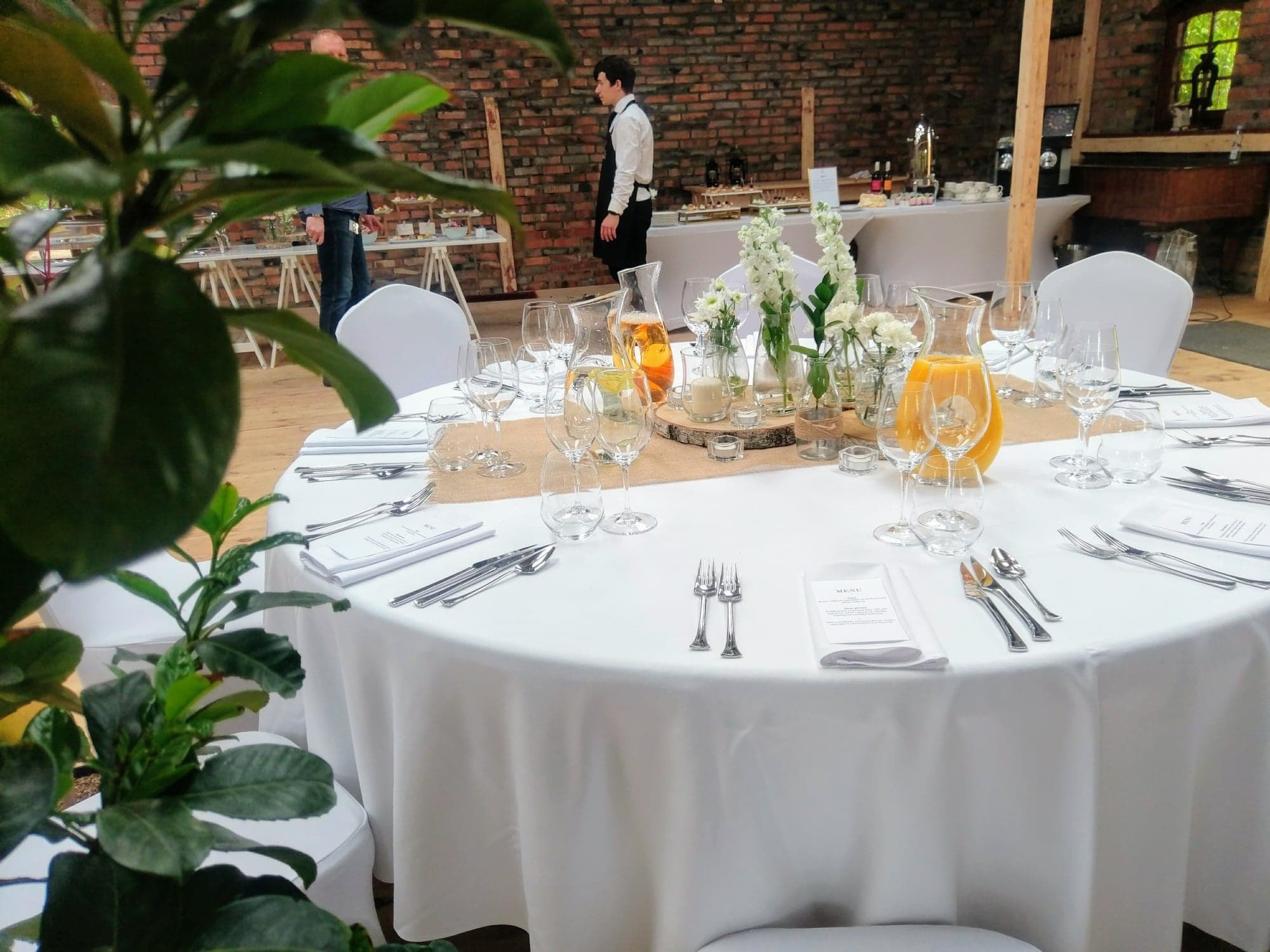 Uroczysty stół z białym obrusem, nakryty zastawą, dzbankami z sokiem i dekoracjami kwiatowymi, ustawiony w rustykalnym wnętrzu z ceglaną ścianą i widocznym personelem w tle.