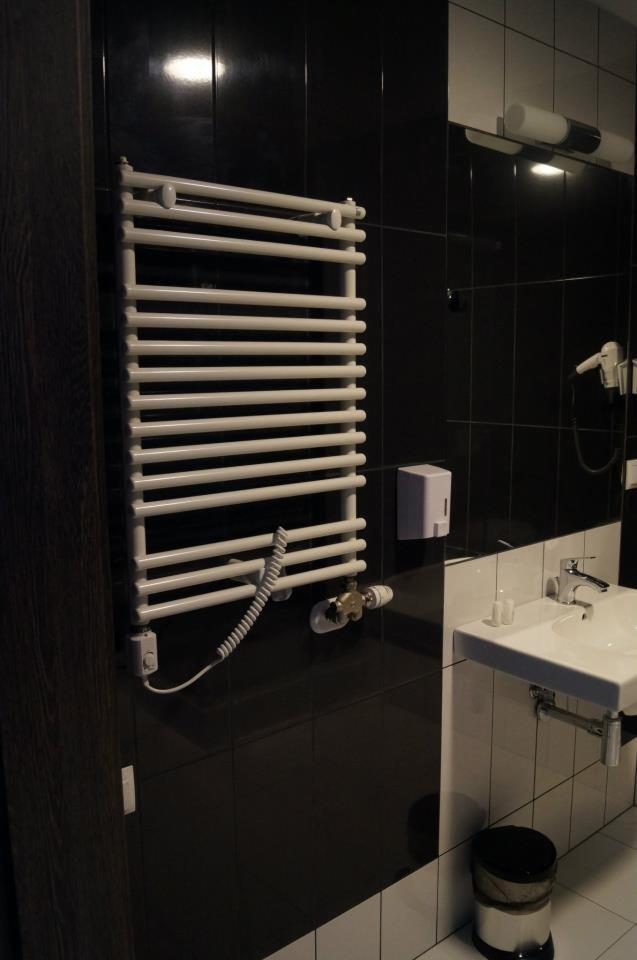Biały, elektryczny grzejnik drabinkowy zamontowany na czarnej ścianie w łazience z białą umywalką i lustrem.