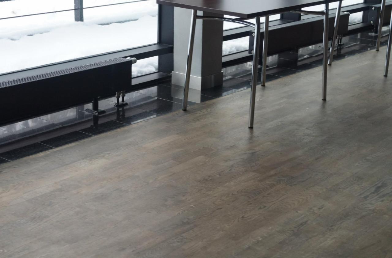 Czarny grzejnik panelowy zamontowany pod oknem z widokiem na śnieg, na tle ciemnej, błyszczącej podłogi i drewnianej podłogi.