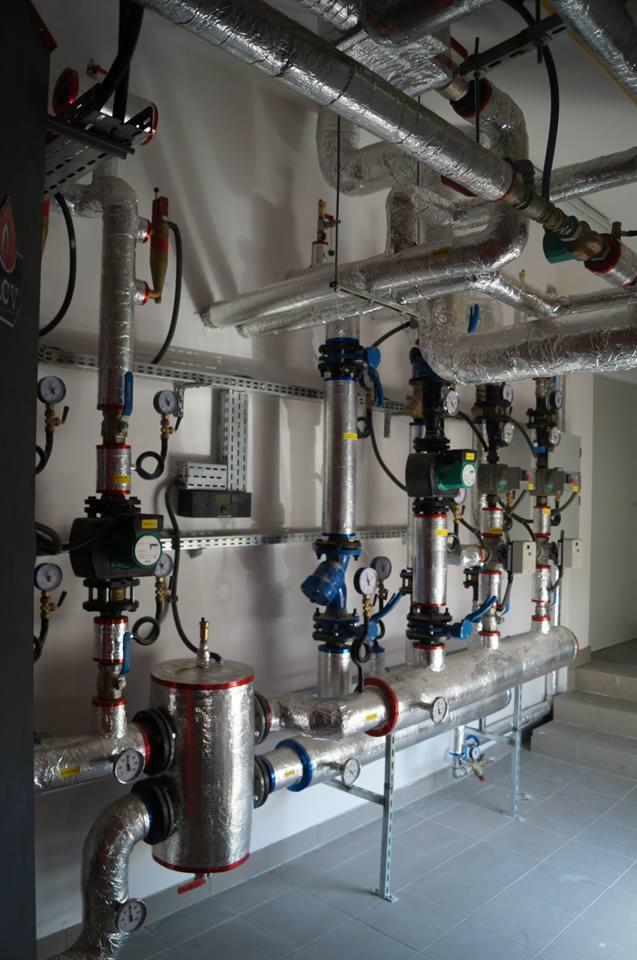 Instalacja centralnego ogrzewania z rurami izolowanymi folią aluminiową, zaworami i manometrami, widok na kotłownię.