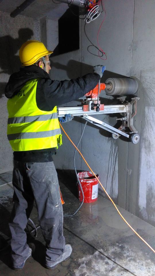 Pracownik w żółtym kasku i kamizelce wierci otwór w betonowej ścianie za pomocą wiertnicy diamentowej, z widocznym czerwonym wiadrem i przewodami elektrycznymi.