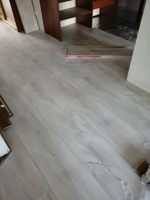 Jasne panele podłogowe z widoczną fakturą drewna, ułożone w pokoju z brązowymi meblami, paczka paneli w folii w tle.