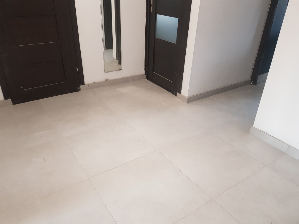 Jasne, kwadratowe płytki podłogowe, ciemne, minimalistyczne drzwi wewnętrzne z ościeżnicami, listwy przypodłogowe w odcieniach szarości.