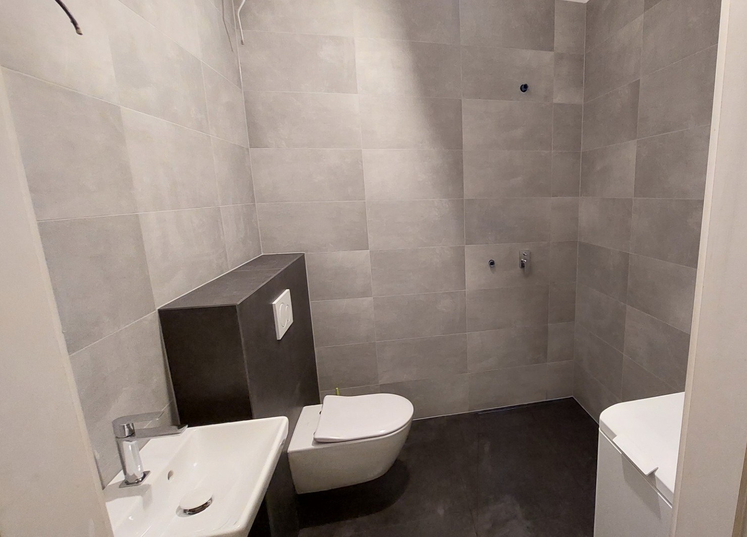 Nowoczesna łazienka z szarymi płytkami, wiszącą toaletą i umywalką. Widoczne niedokończone elementy instalacji wodnej na ścianie.