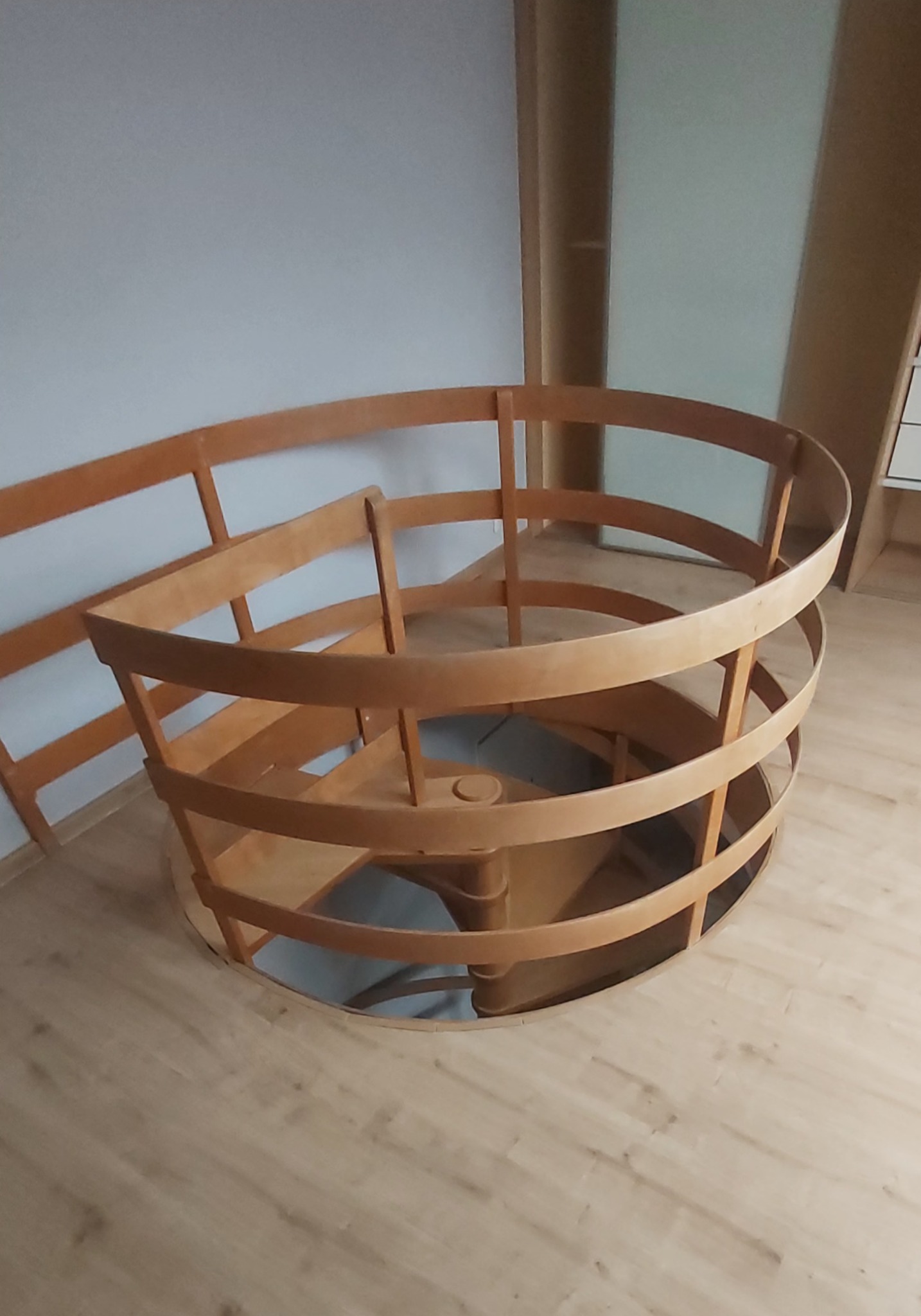 Drewniane schody spiralne z jasnego drewna, widoczne z góry, z zamontowanymi barierkami, na tle jasnej ściany i podłogi z paneli.