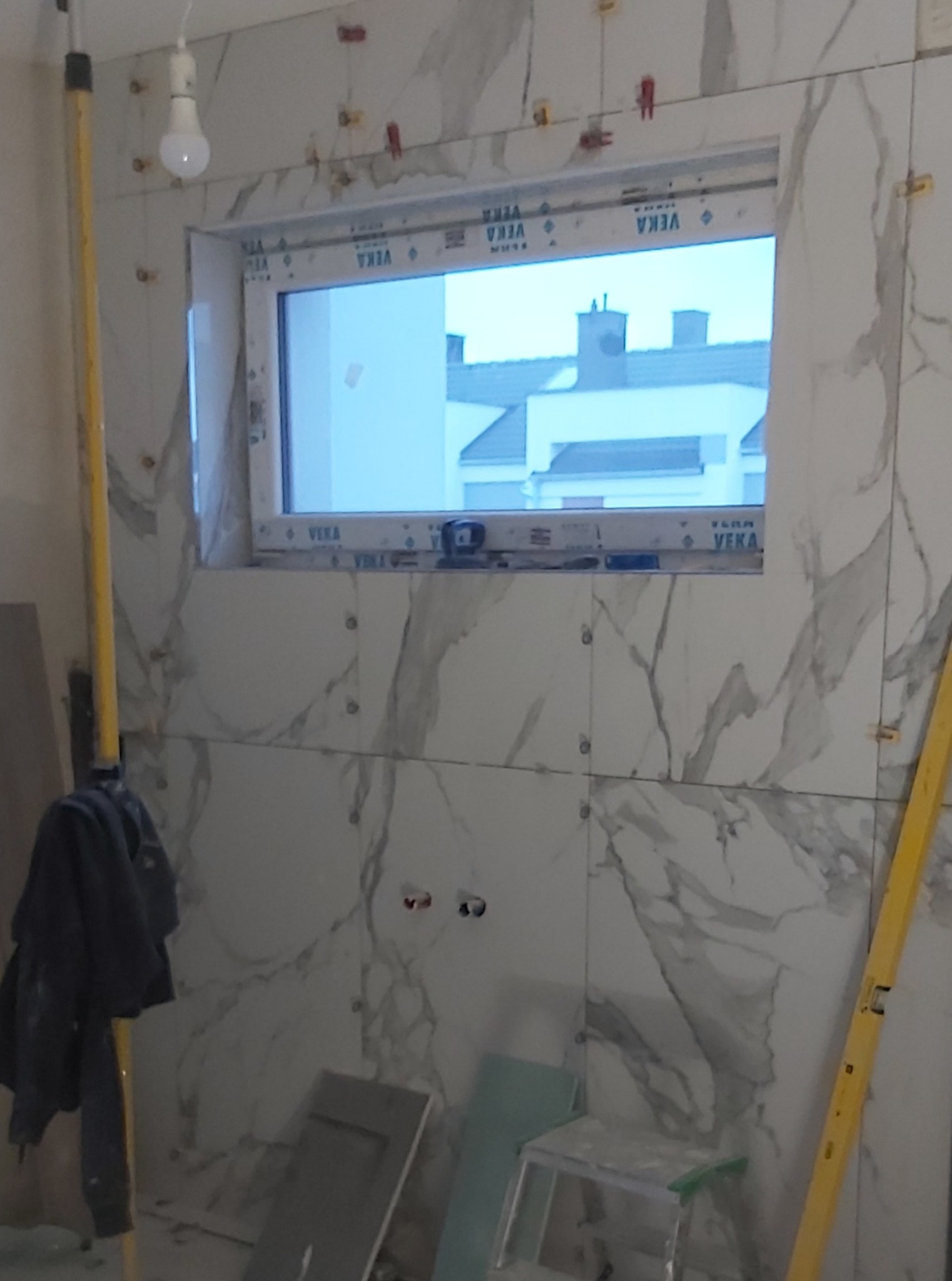 Układanie płytek imitujących marmur wokół okna, z użyciem krzyżyków dystansowych i poziomicy, widoczne niezabudowane gniazdka elektryczne i lampa wisząca