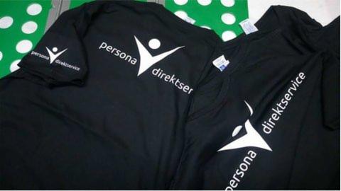 Czarne koszulki z białym nadrukiem logo 'persona direktser', leżące na zielonym materiale w białe grochy.