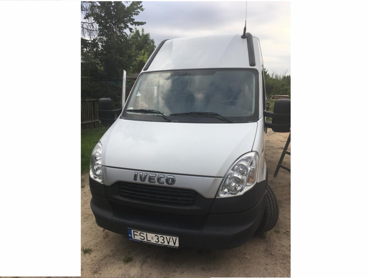 Biały bus Iveco z anteną na dachu, zaparkowany na żwirowym podłożu, z widoczną tablicą rejestracyjną FSL 33VV i odbiciem w przedniej szybie.