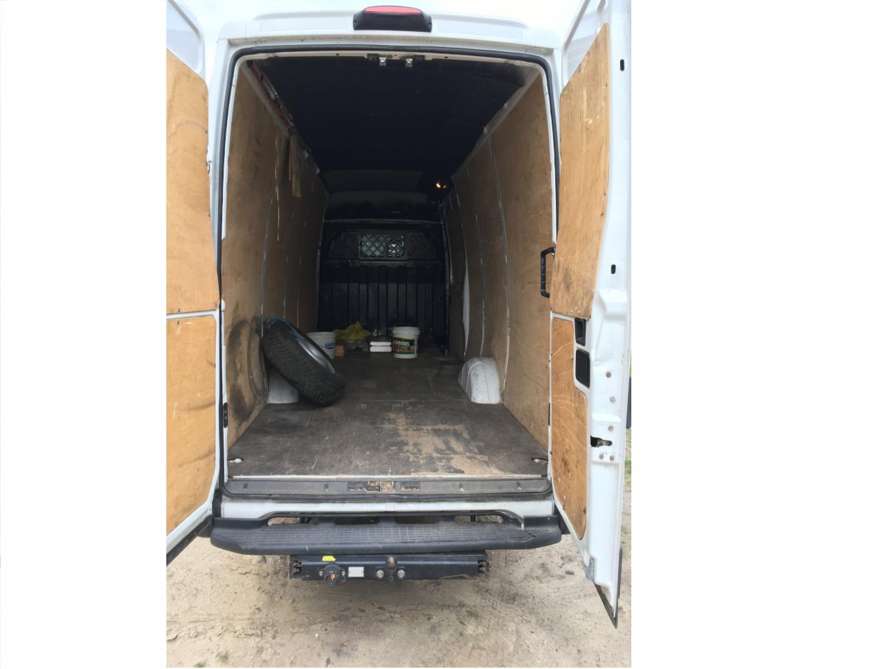 Wnętrze białego furgonetki z otwartymi tylnymi drzwiami, wyłożone sklejką, widoczne opona i wiaderko z farbą w przestrzeni ładunkowej.