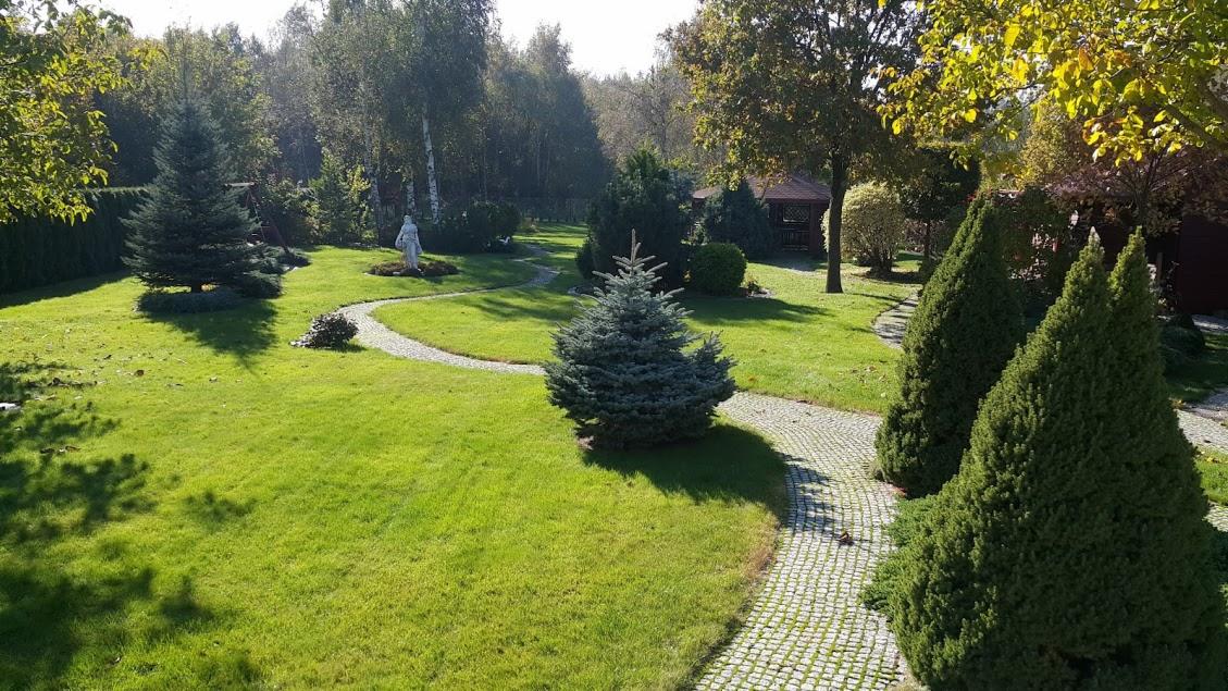 Widok na zadbany ogród z równo przystrzyżonym trawnikiem, ścieżką z kostki brukowej, ozdobnymi krzewami iglastymi i rzeźbą w tle.