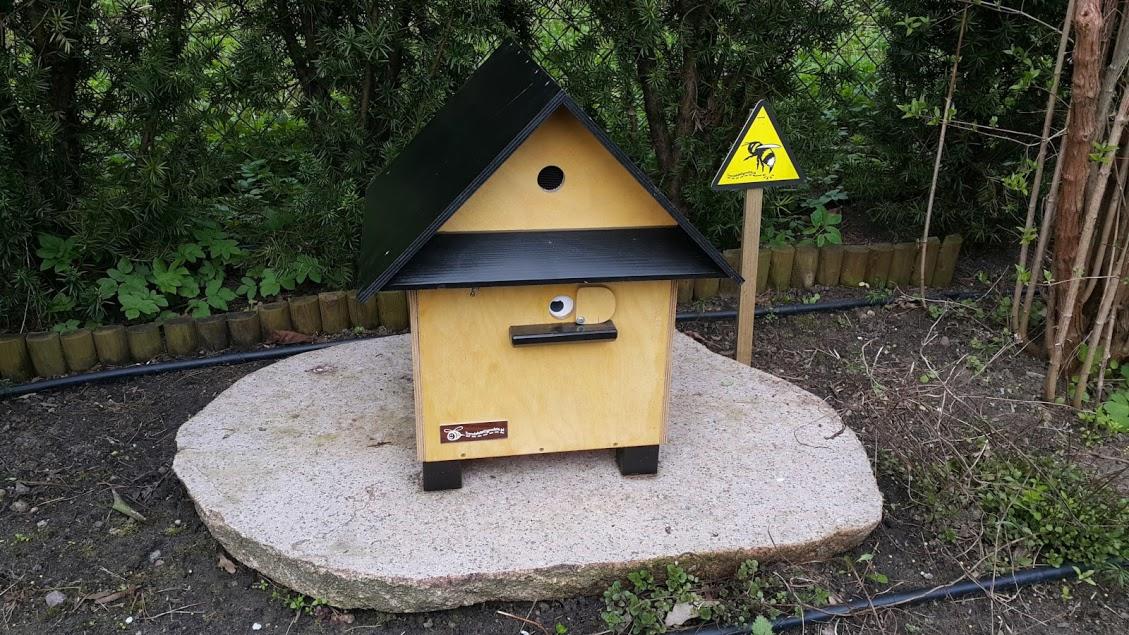 Drewniany domek dla owadów zapylających z czarnym dachem, umieszczony na kamiennej podstawie w ogrodzie, obok tabliczka ostrzegawcza z symbolem pszczoły.
