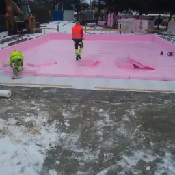 Montaż izolacji termicznej z różowych płyt styropianowych na fundamencie budynku w zimowych warunkach, widoczni dwaj pracownicy w odblaskowych ubraniach, jeden w trakcie pracy z nożem.