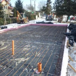 Przygotowany zbrojony szkielet fundamentu na czarnej folii, otoczony różowym styropianem, widoczne wystające rury kanalizacyjne, śnieg na obrzeżach wykopu, w tle koparka i zabudowa jednorodzinna.