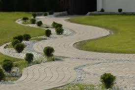 Kręta ścieżka z jasnej kostki brukowej, wyłożona białym grysem i obsadzona kulistymi krzewami na tle zielonego trawnika.