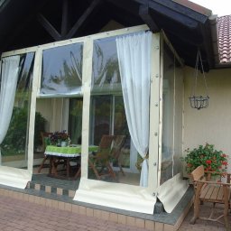 Olplan- plandeki, produkcja i naprawa - Zadaszenie tarasu z przezroczystych pokrowców foliowych z beżowymi obramowaniami, częściowo zasłonięte białymi zasłonami, z widocznym stołem i krzesłami wewnątrz.