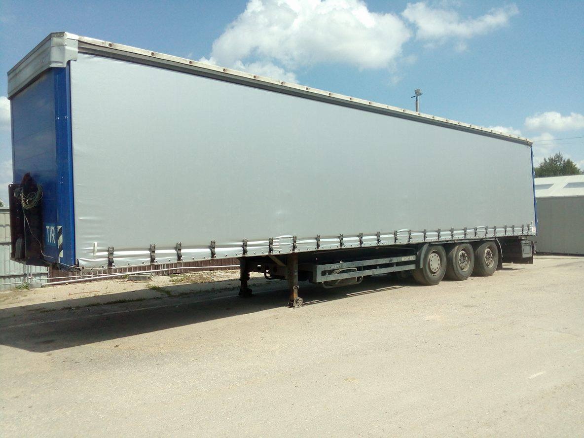 Naczepa TIR z szarą plandeką na parkingu w słoneczny dzień, widoczne mocowania i koła.