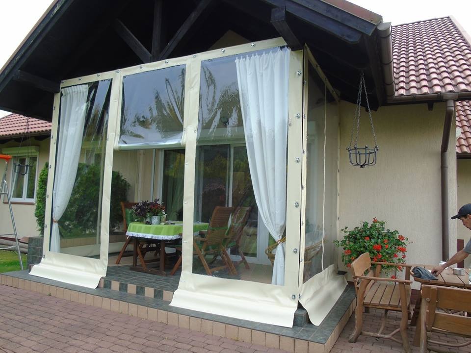 Zadaszenie tarasu z przezroczystych pokrowców foliowych z beżowymi obramowaniami, częściowo zasłonięte białymi zasłonami, z widocznym stołem i krzesłami wewnątrz.