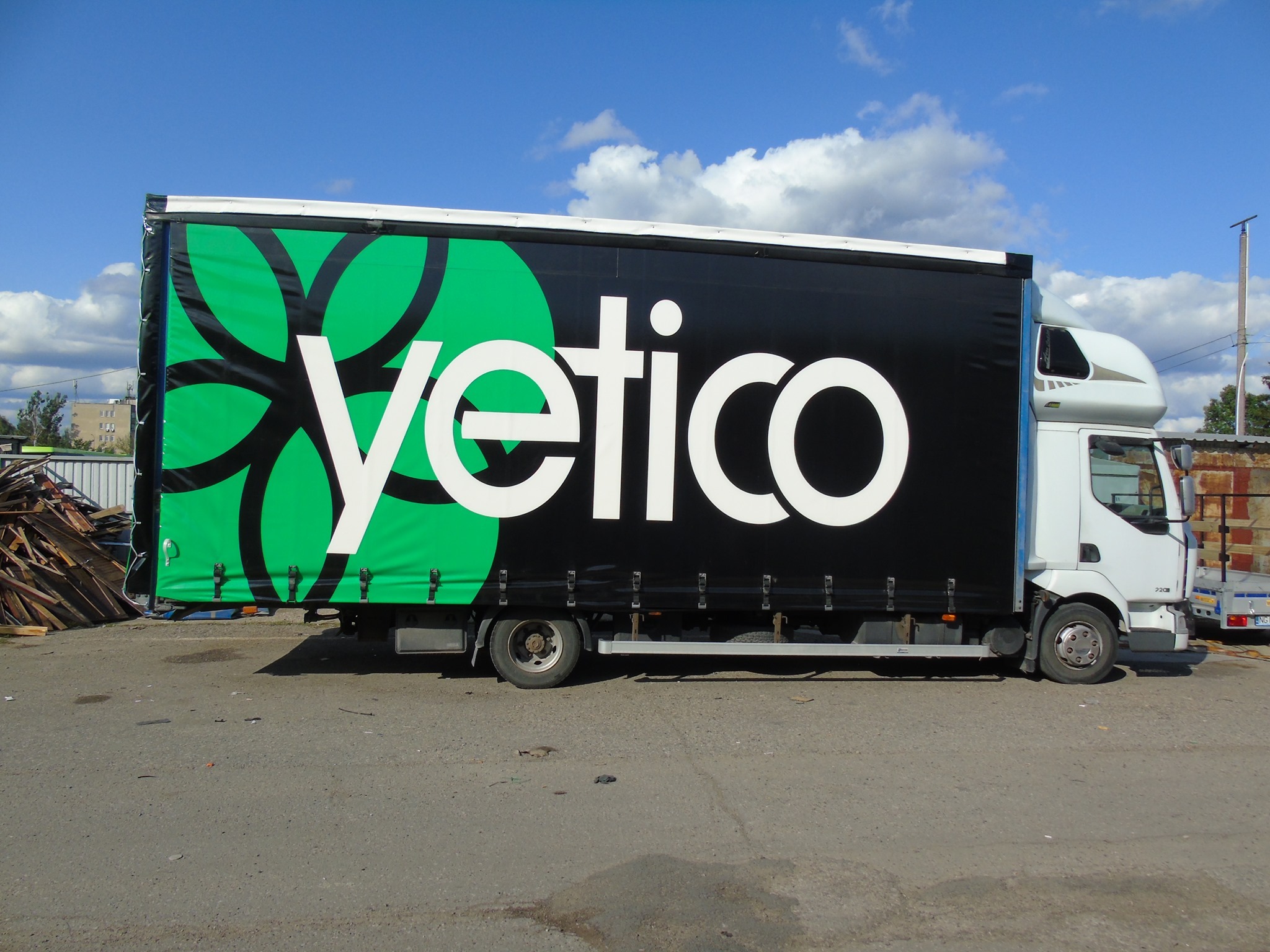 Ciężarówka z czarną plandeką ozdobioną dużym, zielonym, abstrakcyjnym motywem roślinnym i białym logo 'yetico' zaparkowana na szarym asfalcie pod błękitnym niebem z chmurami.