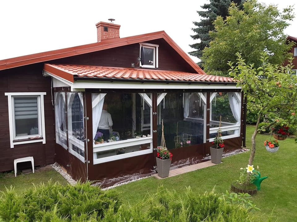 Zadaszony taras z brązowymi, przezroczystymi osłonami, białymi ramami i zasłonami, widok z zewnątrz na tle ogrodu z drzewami i kwiatami.