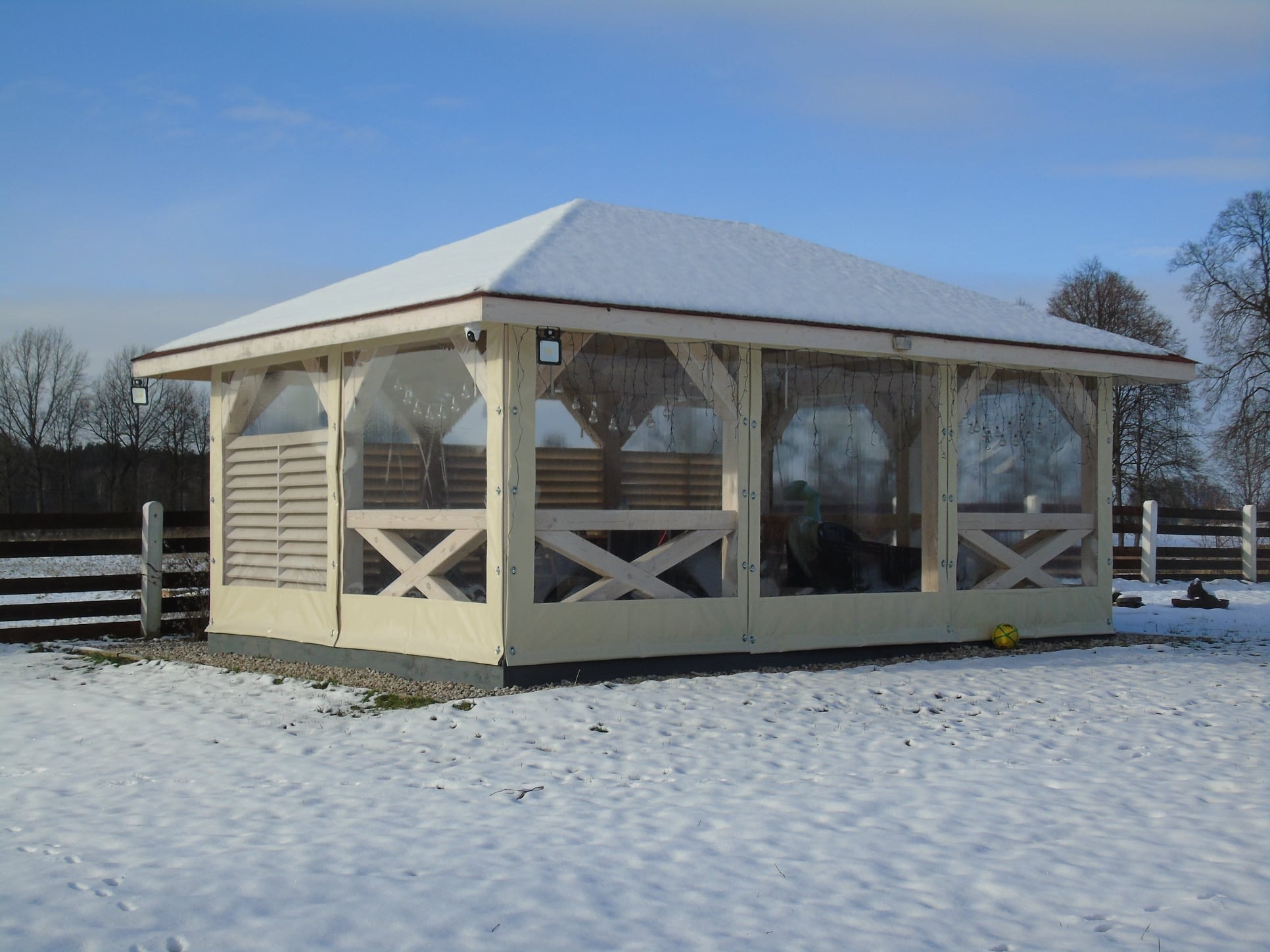 Zadaszona altana z transparentnymi ścianami i śniegiem na dachu, otoczona zimowym krajobrazem.