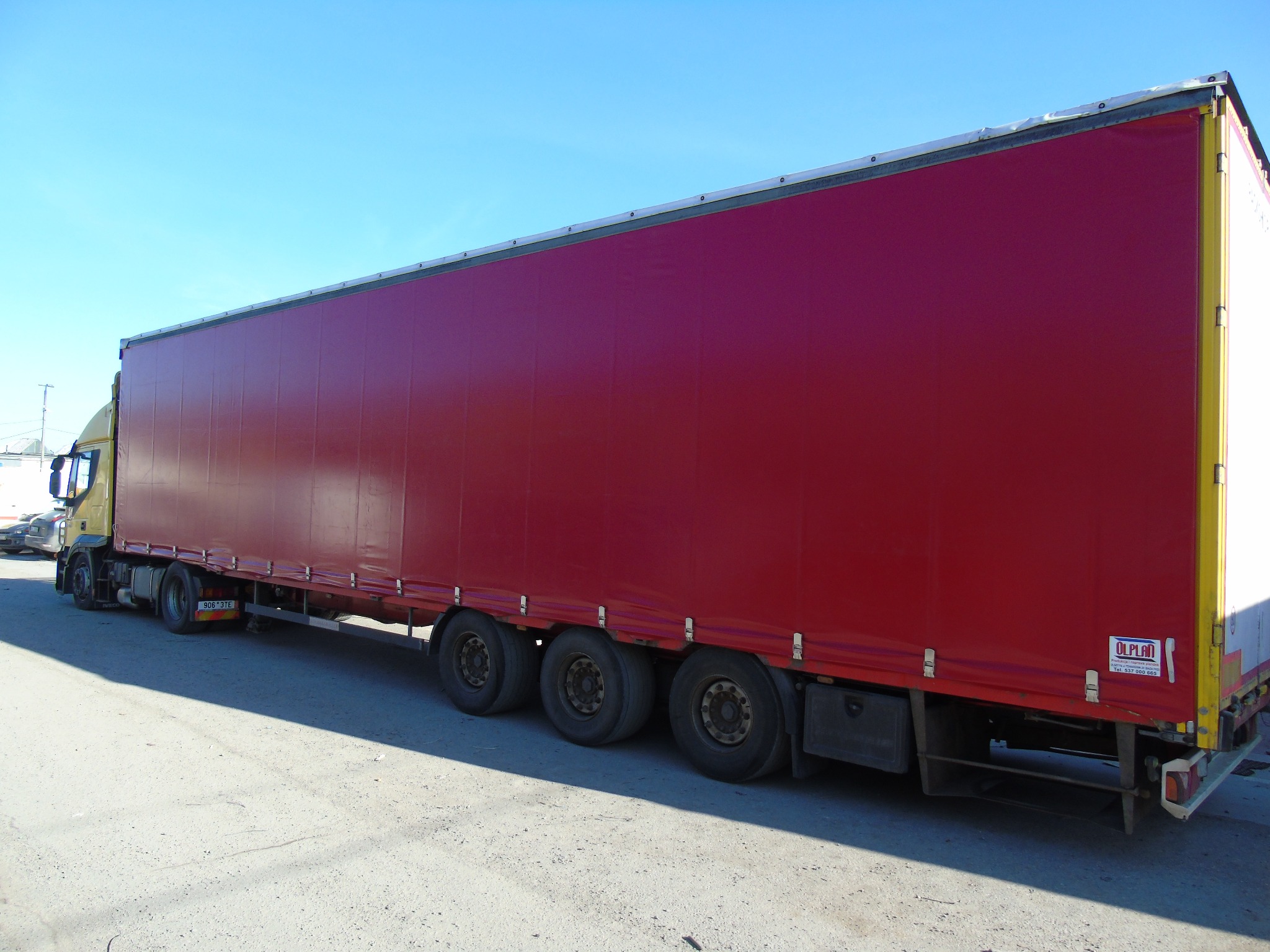 Czerwona plandeka naczepy ciężarówki z widocznymi mocowaniami i logo producenta, na tle błękitnego nieba.