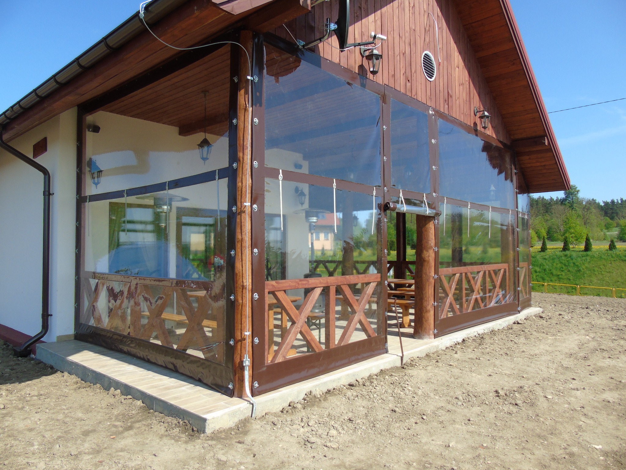 Zewnętrzna osłona z przezroczystej folii PVC na drewnianej konstrukcji tarasu, zamocowana za pomocą metalowych oczek i linek, widoczne meble ogrodowe wewnątrz.