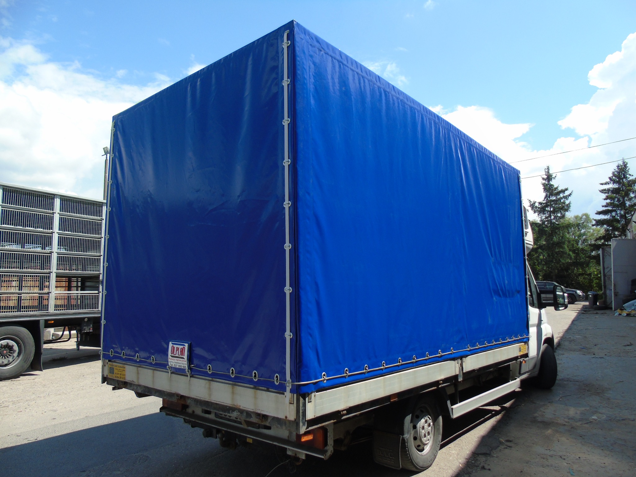 Niebieska plandeka na samochodzie dostawczym, widok z tyłu, z widocznymi elementami mocującymi i logo producenta na plandece.